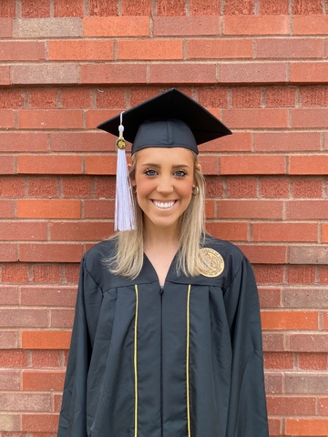 Carina standing in front of a brick wall smiling wearing her University of Iowa cap and gown