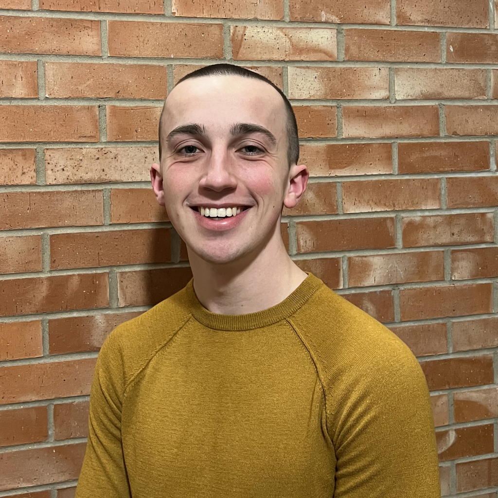 Carson is in a yellow shirt smiling in front of a brick wall